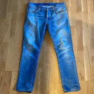 Blå jeans från Nudie - Snygga blå jeans från Nudie med klassisk femficksdesign och orange sömmar. De har en straight passform och är tillverkade i slitstarkt denim. Perfekta för en avslappnad stil. Passar både avslappnade och finare tillfällen