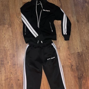 Svart träningsset från Palm Angels - Snyggt svart träningsset från Palm Angels med vita ränder längs ärmar och ben. Jackan har dragkedja och två fickor med vita detaljer. Perfekt för en sportig look eller avslappnad stil. Storlek i M och den är mycket sparsamt använd🖤