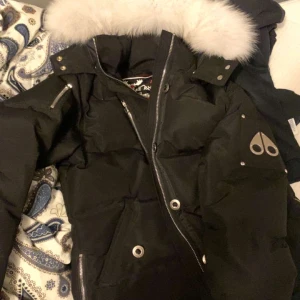 Svart dunjacka från Moose Knuckles - Säljer en svart dunjacka från Moose Knuckles med en lyxig pälsdetalj på huvan. Jackan har en snygg design med dragkedja och flera fickor med knappar. Perfekt för kalla dagar.