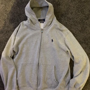 Grå hoodie från Ralph Lauren - Säljer en stilren grå hoodie från Ralph Lauren med dragkedja och klassisk logga på bröstet. Perfekt för en avslappnad look. Hoodien har långa ärmar och en bekväm passform. Medföljer snören till luvan, priset kan diskuteras 