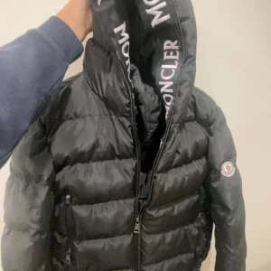 Svart pufferjacka från Moncler - Säljer en snygg svart pufferjacka från Moncler med broderad logga på ärmen. Jackan har en hög krage och dragkedja framtill. Perfekt för kyliga dagar med sin stilrena design och varma material.