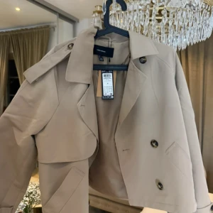 Beige trenchcoat från Vero Moda - Säljer en stilren beige trenchcoat från Vero Moda. Jackan har en klassisk design med knappar framtill och en elegant krage. Perfekt för att ge en sofistikerad touch till din outfit. Aldrig använd 