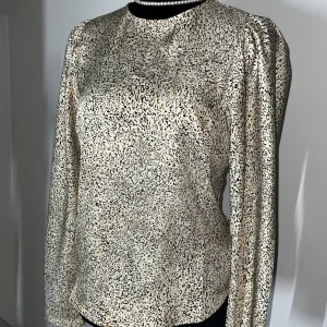 Mönstrad blus med långa ärmar - Snygg blus med leopardmönster i svart och beige. Blusen har långa ärmar och en dragkedja i ryggen för enkel påklädning. Perfekt för att ge en stilren touch till din outfit.