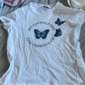 Vit t-shirt med fjärilar från New yorker - Säljer en vit t-shirt från New yorker med tryck av blå fjärilar och texten 'Set Yourself Free'. T-shirten har korta ärmar och en rund halsringning. Perfekt för en avslappnad stil.