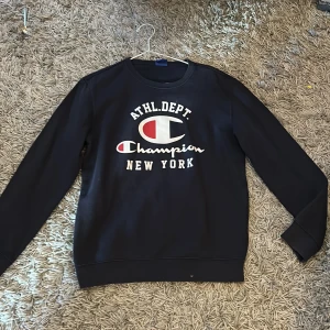 Svart sweatshirt från Champion - Säljer en svart sweatshirt från Champion med tryck 'ATHL.DEPT. Champion NEW YORK' på framsidan. Tröjan har långa ärmar och en klassisk rund halsringning. Perfekt för en avslappnad stil. Passar perfekt med t.ex en piké under. Pris är diskuterbart.