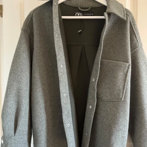 Grå overshirt från Zara - Säljer en stilren grå overshirt från Zara. Den har en klassisk krage och knäppning framtill med vita knappar. Perfekt för lager-på-lager-stil. En bröstficka ger en extra touch av funktionalitet.