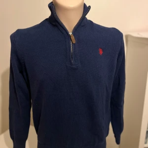 Mörkblå tröja med dragkedja från U.S. Polo Assn. - Säljer en stilren mörkblå tröja från U.S. Polo Assn. med dragkedja och broderad logga på bröstet. Tröjan har långa ärmar och en hög krage, perfekt för kyligare dagar. Passar både till vardags och mer uppklädda tillfällen.