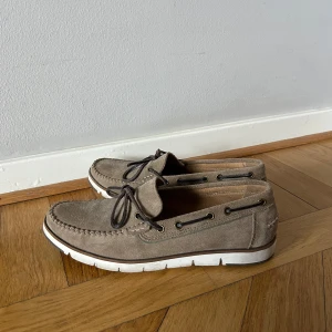 Beige mockaloafers från Geox - Snygga beige loafers från Geox i mocka med vit sula och snörning. Perfekta för en stilren look. Skorna har en klassisk design med dekorativa sömmar och är bekväma att bära.