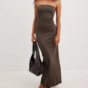 Brun långklänning från NA-KD - Säljer en elegant brun långklänning från NA-KD i storlek XS. Klänningen har en stilren design med en slits som ger en snygg detalj. Perfekt för en chic look! Slit bakom klänningen för att kunna röra på sig. Använd en gång. 