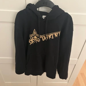 Svart hoodie från Saint Laurent - Säljer en svart hoodie från Saint Laurent med en cool stjärndesign och text i guld på framsidan. Tröjan har en klassisk passform med långa ärmar och en praktisk ficka framtill. Perfekt för en stilren och avslappnad look.