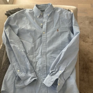 Ljusblå skjorta från Ralph Lauren - Snygg ljusblå skjorta från Ralph Lauren med klassisk krage och broderad logga på bröstet. Skjortan har långa ärmar och knappar framtill. Perfekt för en stilren look. Oanvänd