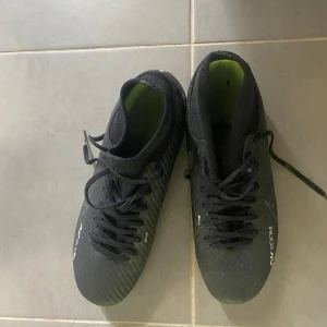 Svarta Nike fotbollsskor med gula detaljer - Säljer ett par svarta Nike fotbollsskor med gula detaljer och snörning. Skorna har en modern design med Zoom-teknologi för extra komfort och prestanda på planen. Perfekta för fotbollsspelare som vill ha stil och funktionalitet.