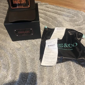 Svart keps med broderad text - Snygg svart keps från John Hatter & Co med broderad text 'THUG LIFE' i orange på framsidan. Perfekt för en cool och avslappnad  stil. Kommer med originalförpackning och påse. Har kvar kvittot, jag köpte den från season man pris kan diskuteras!