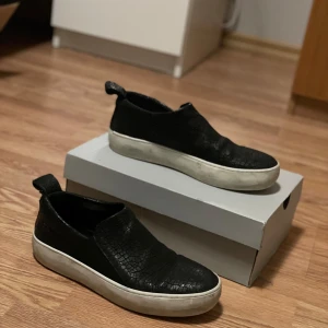 The last conspiracy  - Snygga svarta sneakers med en vit sula och ett krackelerat mönster på ovandelen. Skorna har en praktisk dragflik baktill för enkel påtagning. Perfekta för en stilren look. Alligator skin. En bit har lossnat på bak sidan om skon. Därav priset. Fler frågor eller funderingar är det bara att höra av er😁