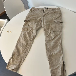 Beige cargopants med dragkedja - Snygga beige cargopants från Stayhard med flera fickor och dragkedja vid bensluten. Perfekta för en avslappnad stil med praktiska detaljer. Passar bra till sneakers eller kängor.