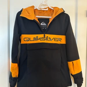 Svart och orange jacka från Quiksilver - Säljer en fodrad svart och orange anorak/jacka från Quiksilver. Jackan har en halvlång dragkedja framtill och en stor Quiksilver-logga över bröstet. Den har långa ärmar, muddar med tumhål , stor ficka vid magen och en praktisk huva. Perfekt för höst- och vinterdagar. Storleken är barn M/12år, ca xxs eller strl 152. Vind-/vattentät med 10 000mm vattenpelare med Quicksilver Dry Light Technology. 