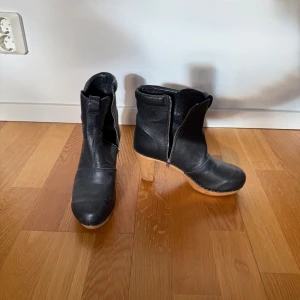 Svarta stövletter i skinn med klack och dragkedja - Snygga svarta stövletter i skinn med träklack och dragkedja på sidan. Perfekta för en stilren look. De har en rund tå och en bekväm passform. Endast använda 2 gånger så i jättefint skick. Det är en större 38 då jag normalt brukar ha 39.