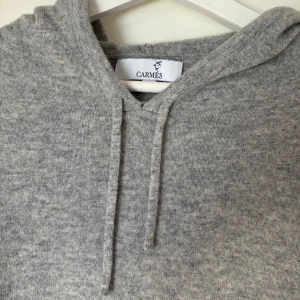 Grå kashmir hoodie från Carmes - Säljer en supermjuk och stilren grå hoodie från Carmes, tillverkad i 100% kashmir. Perfekt för kyliga dagar med sin bekväma passform och klassiska design. Den har en justerbar huva med dragsko och ribbade muddar vid ärmslut och nederkant. Ett måste för garderoben! 