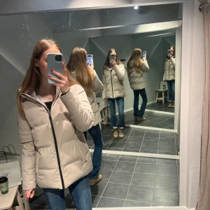 Beige pufferjacka med dragkedja - Säljer en snygg beige pufferjacka med dragkedja. Jackan har en stilren design och är perfekt för kyligare dagar. Den har en hög krage och långa ärmar för extra värme. Passar bra till jeans och boots.