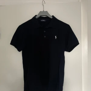 Mörkblå  pikétröja från Ralph Lauren - Snygg mörkblå pikétröja från Ralph Lauren med klassisk krage och knappar. Den har en broderad logga på bröstet i vitt. Perfekt för en stilren look. Skick 8/10 