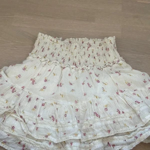 Blommig kjol från Neo Noir - Charmig blommig kjol från Neo Noir i en mjuk vit nyans med små rosa och gula blommor. Kjolen har en volangdetalj och en elastisk midja för en bekväm passform. Perfekt för en somrig look!