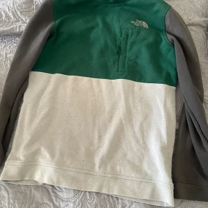 Grön och beige långärmad tröja från The North Face - Säljer en snygg långärmad tröja från The North Face i grönt och beige med grå ärmar. Tröjan har en praktisk bröstficka med dragkedja och är perfekt för kyligare dagar. Passar bra till både vardag och äventyr.