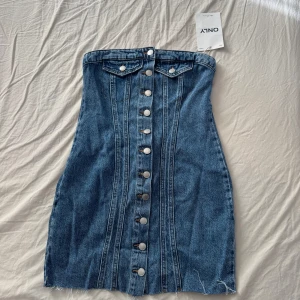 Jeansklänning från ONLY - Snygg axelbandslös jeansklänning från ONLY i blå denim. Klänningen har knappar framtill och en smickrande passform med korsettliknande sömmar. 