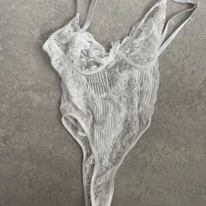 Vit spetsbody - Elegant vit spetsbody med justerbara axelband och vacker blommig spetsdetalj. Den har en djup urringning och en liten rosett framtill. Perfekt för en speciell kväll.