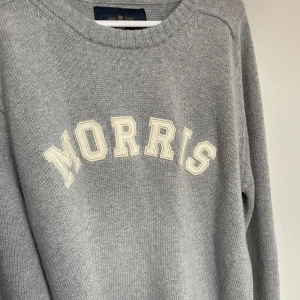Grå stickad tröja från Morris - Säljer en stilren grå stickad tröja från Morris med texten 'Morris' i vitt på framsidan. Tröjan har långa ärmar och en rund halsringning. Perfekt för en avslappnad och snygg look.