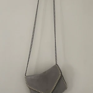 Grå axelväska med kedjedetaljer - Snygg grå axelväska med coola kedjedetaljer längs kanterna. Väskan har en praktisk dragkedja och är perfekt för att bära dina nödvändigheter. Den har en stilren design som passar till många olika outfits.