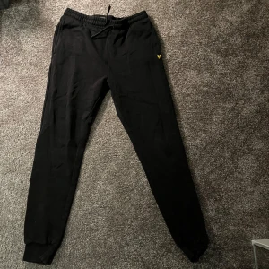 Svarta mjukisbyxor från Lyle & Scott - Säljer ett par svarta mjukisbyxor från Lyle & Scott med elastisk midja och snörning. Byxorna har en broderad logga i gult på sidan. Perfekta för en avslappnad stil. Använd 1 gång 