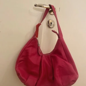 Röd axelväska från Rizzo - Snygg röd axelväska från Rizzo i skinn. Väskan har en dragkedja upptill och en enkel axelrem. Perfekt för att ge en färgklick till din outfit💕
