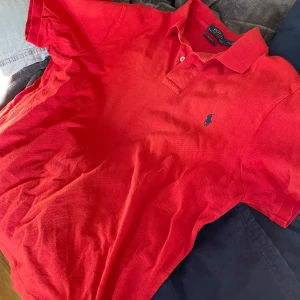 Röd pikétröja från Ralph Lauren - Säljer en snygg röd pikétröja från Ralph Lauren i slim fit. Tröjan har korta ärmar och en klassisk krage med knappar. Den ikoniska loggan är broderad på bröstet. Perfekt för en stilren look! Helt ny använd en gång