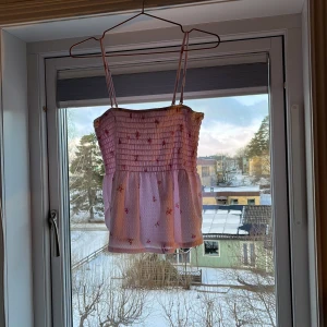 Rosa blommig linnetopp - Söt rosa linnetopp med blommigt mönster och smala axelband. Toppen har en smockad överdel som ger en fin passform och en luftig nederdel. Perfekt för varma sommardagar!