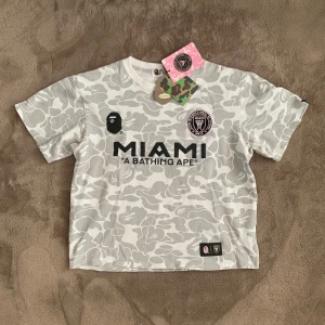 Vit/Grå Inter Miami x Bape T-Shirt - Säljer en Inter Miami x Bape T-shirt med tags. Det står strl L men den passar mer som en XS då den har krympt i tvätten, därav priset också. Vid frågor eller funderingar skicka ett pm, Mvh