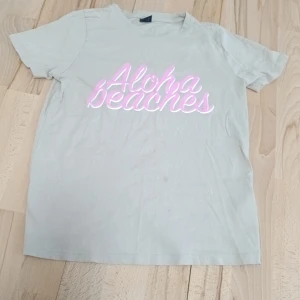 Beige t-shirt från Gina Tricot - Säljer en beige t-shirt från Gina Tricot med texten 'Aloha Beaches' i rosa på framsidan. Perfekt för en avslappnad stil. Kortärmad och i mjukt material.