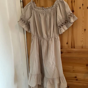 Beige klänning med spetsdetaljer - Charmig beige klänning med offshoulder-design och spetsdetaljer på ärmarna. Klänningen har en volangkant nedtill och elastisk midja för en bekväm passform. Perfekt för en somrig look!