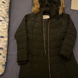 Svart dunjacka med fuskpäls - Säljer en svart dunjacka från Vila med fuskpäls på huvan. Jackan har en quiltad design och dragkedja framtill. Perfekt för kyliga dagar.