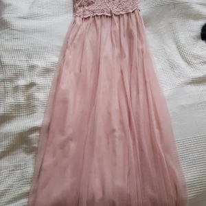 Gammelrosa fiklänningen med spetsliv - Elegant rosa långklänning med spetsdetaljer på överdelen och ett luftigt, flödande kjolparti. Klänningen har en vacker öppen rygg och är ärmlös, vilket ger en romantisk och sofistikerad look. Perfekt för speciella tillfällen. Elastiskt tyg,insydd i midjan då passformen tidigare var lös och stor i storlek. Använd på bröllop en gång.