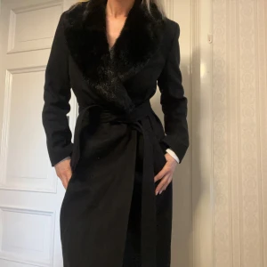 Svart kappa med pälsdetalj - Elegant svart kappa med en lyxig pälsdetalj vid kragen. Kappan har en knytning i midjan som ger en snygg siluett. Perfekt för kyligare dagar när du vill känna dig både varm och stilfull.