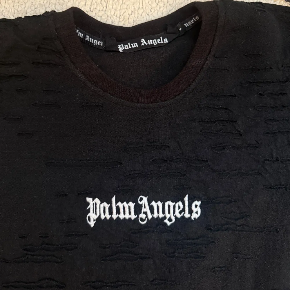 Säljer en svart tröja från Palm Angels med ett unikt texturerat mönster. Tröjan har långärmade ärmar och Palm Angels-loggan i vitt på både framsidan och baksidan. Perfekt för en stilren och modern look.. Neuletakit & Villapaidat.