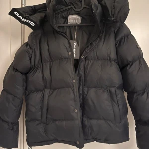 Svart pufferjacka från Gabos - Säljer en svart pufferjacka från Gabos i mycket bra skick. Jackan är perfekt för vintern med sin tjocka vaddering och långa ärmar. Den har en dragkedja och knappar framtill samt en stor huva för extra värme. Superbekväm och stilren för kalla dagar! ❄️