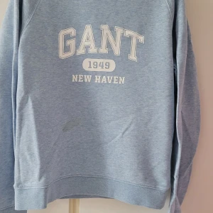 Ljusblå sweatshirt från GANT - Säljer en snygg ljusblå sweatshirt från GANT med tryck 'GANT 1949 NEW HAVEN' på framsidan. Tröjan är i bra skick och perfekt för en avslappnad stil. Den har långa ärmar och en klassisk rund halsringning. Perfekt för både vår och höst!