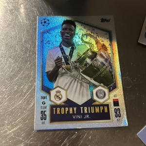 Fotbollskort Trophy Triumph Vini Jr. - Säljer ett coolt fotbollskort från Topps med Vini Jr. i fokus. Kortet har en glittrig yta och visar Vini Jr. med en stor trofé. Perfekt för samlare av fotbollskort eller fans av Real Madrid. Kortet har detaljer som klubbens emblem och spelarens statistik.