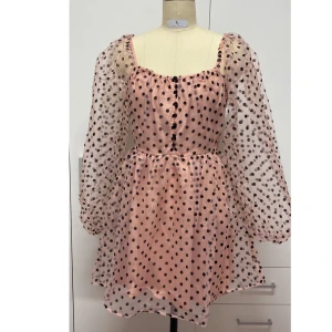 Rosa prickig klänning med puffärmar - Superfin rosa klänning med svarta prickar och puffiga långa ärmar. Den har en söt knappdetalj framtill och är perfekt för vår och sommar. Materialet känns lätt och luftigt, vilket gör den idealisk för varmare dagar. Passar perfekt till både vardag och fest!