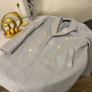 Ljusgrå kappa med guldknappar - Säljer en elegant ljusgrå kappa med dubbelknäppning och stiliga guldknappar. Perfekt för höst och vår, med en klassisk design som passar både till vardags och fest. Kappan har långa ärmar och en snygg krage. Ett måste för garderoben!