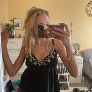 Svart linne med broderade blommor - Säljer ett svart linne med vita broderade blommor längs urringningen. Det har smala axelband och en söt knytning framtill. 