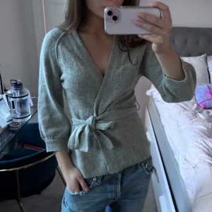 Grå omlottkofta - Säljer en superfin grå omlottkofta med trekvartsärmar. Den har en knytning i midjan med en rosett vilket är så snygg . Perfekt för både vardag och lite finare tillfällen. 