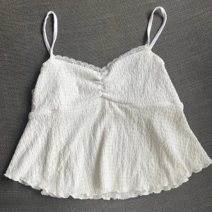 Vit ärmlös topp med spetsdetaljer - Säljer en söt vit topp med tunna axelband och spetsdetaljer längs kanten. Perfekt för sommardagar eller som ett lager under en kofta. Den har en lätt och luftig passform. Sitter inte så bra kvar under bysten om man stäcker upp armarna. Men superfin på verkligen. Strl L men är som en S