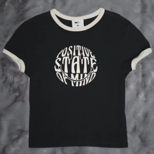 Svart t-shirt med tryck - Mörkgrå t-shirt. Har ett tryck med texten 'Positive State of Mind' i vitt. T-shirten är kortärmad och har vita kanter vid ärmar och hals. Den har en aningen tajtare passform och kortare modell.